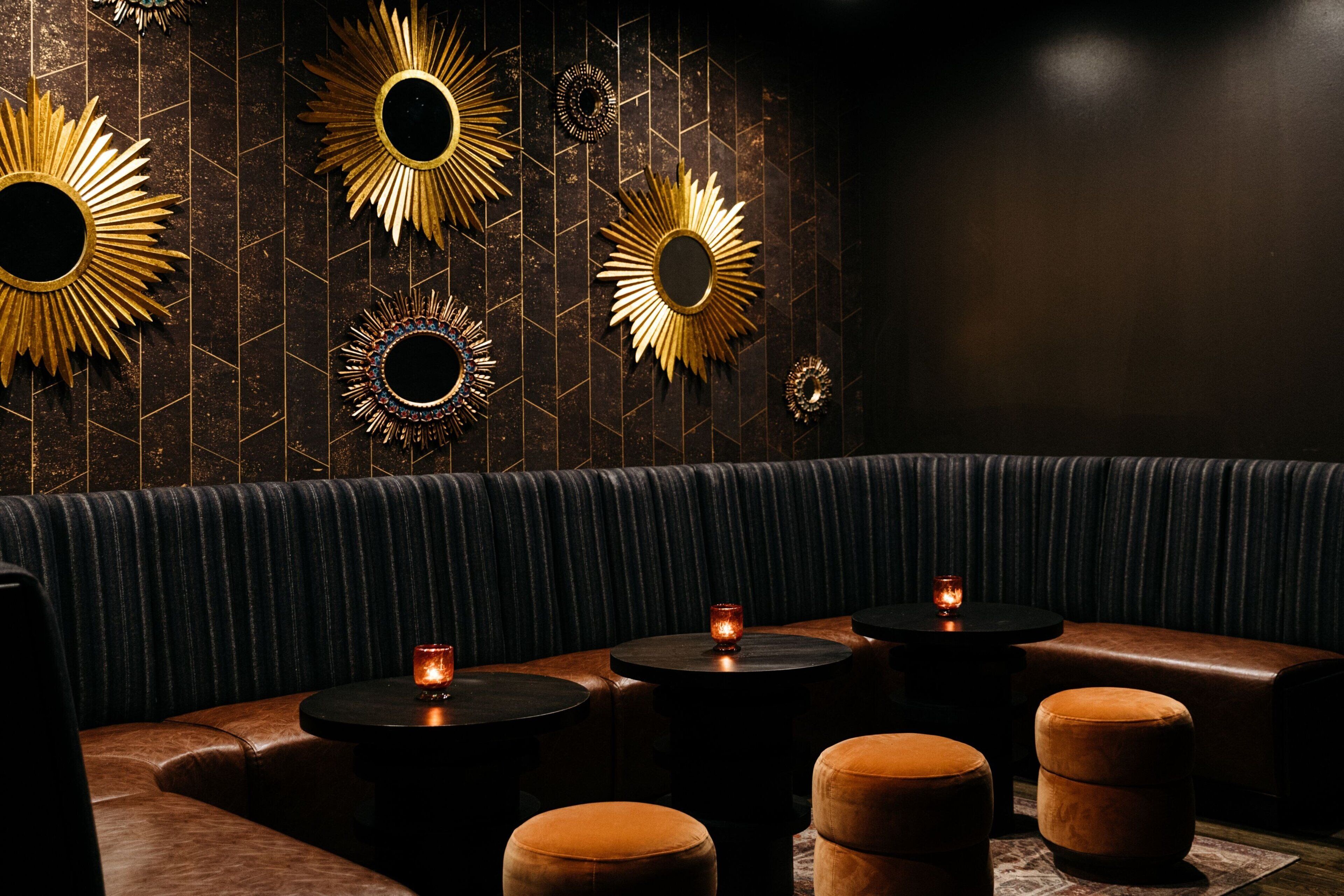 A dimly lit lounge area with leather seating and round tables, featuring wall-mounted decorative mirrors and candles.