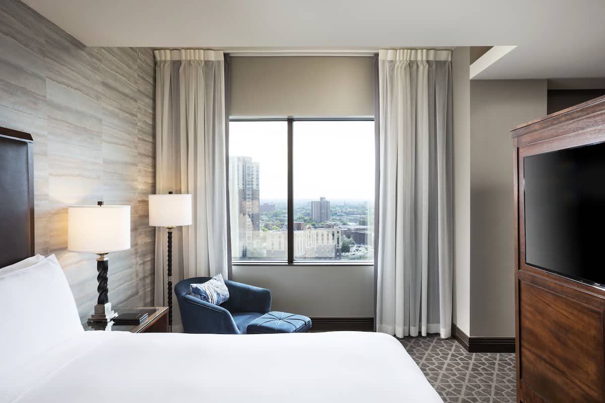 A hotel room with a large bed, a seating area with a chair, a TV, and a window with curtains.