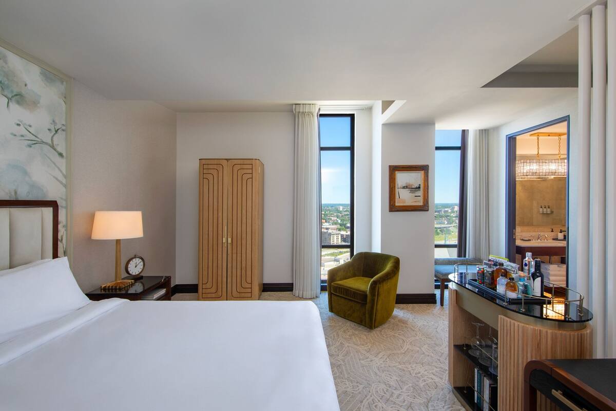 A hotel room with a large bed, a desk with a lamp, a green armchair, a minibar, and a large window with a city view.