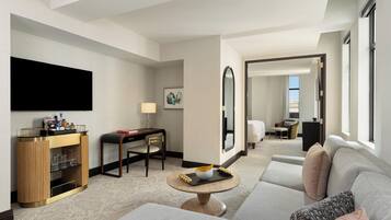 A modern hotel room with a sofa, a round coffee table, a TV, a minibar, and a desk with a chair.