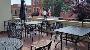 Um terraço com mesas e cadeiras ao ar livre, incluindo mesas redondas e quadradas, e duas bandeiras americanas.