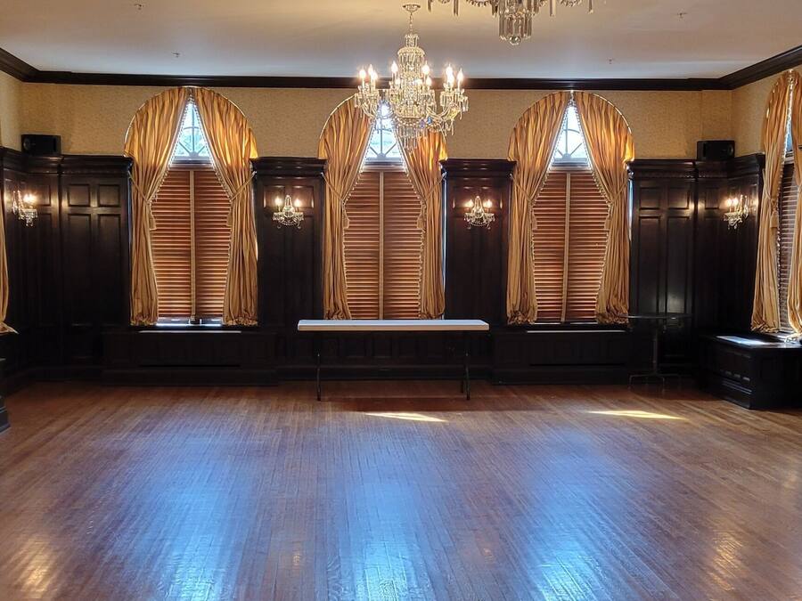 A spacious room with a wooden floor, large windows with curtains, and a chandelier.