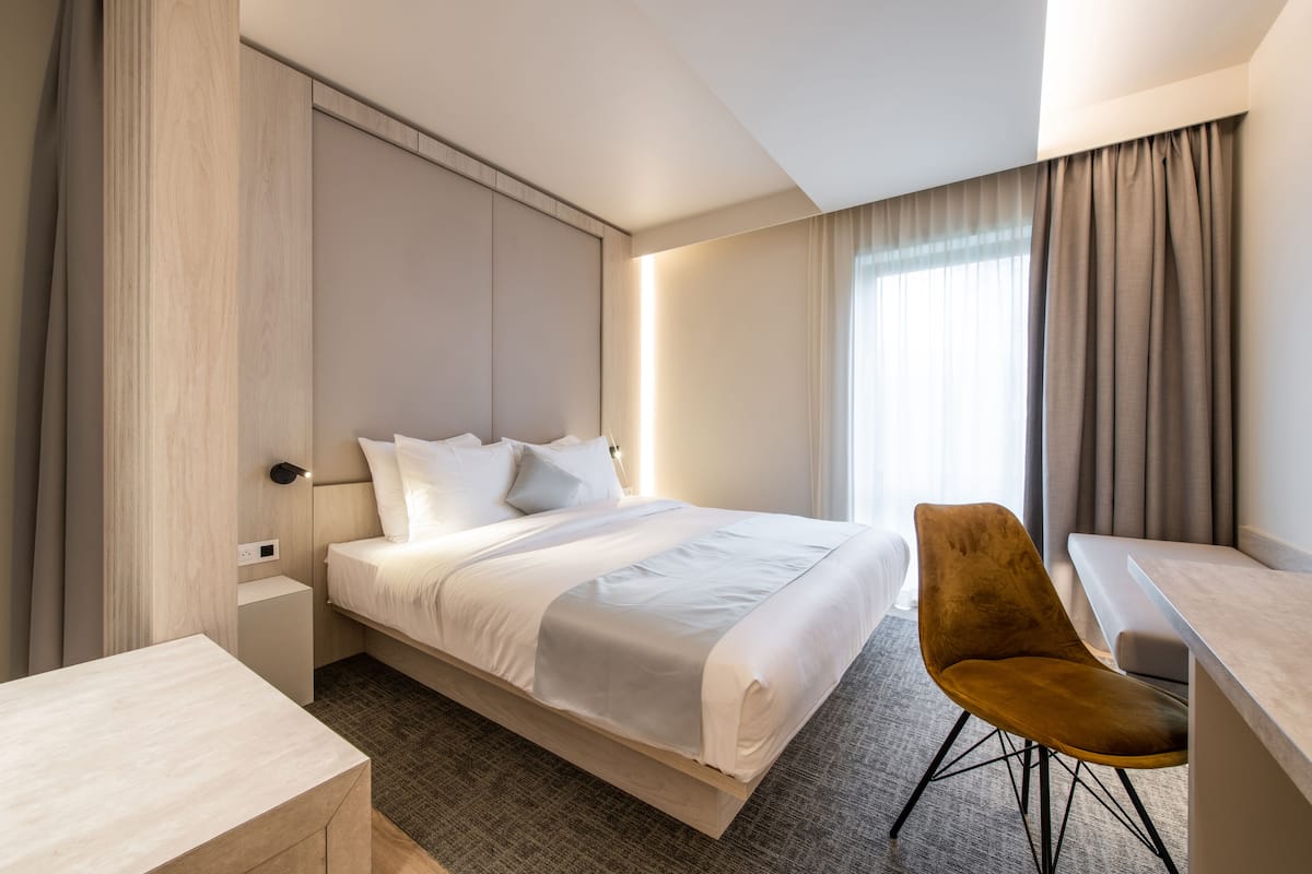 Quarto de hotel moderno com uma cama grande, uma escrivaninha, uma cadeira e uma janela com cortinas.