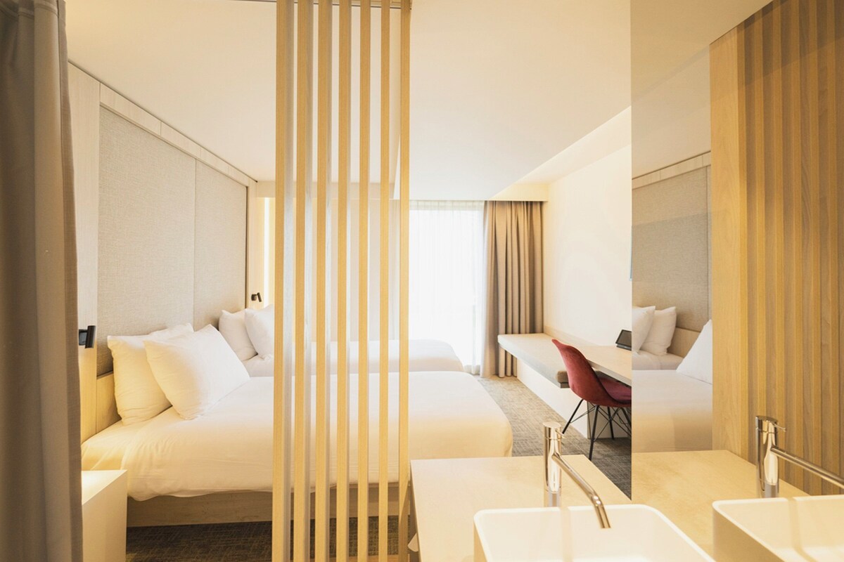 A hotel room with two beds, a desk, a chair, and a bathroom with a sink and mirror.
