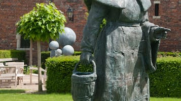 Una estatua de una mujer con un vestido largo sosteniendo una cesta, sobre un pedestal en un césped, con un edificio de ladrillo al fondo.