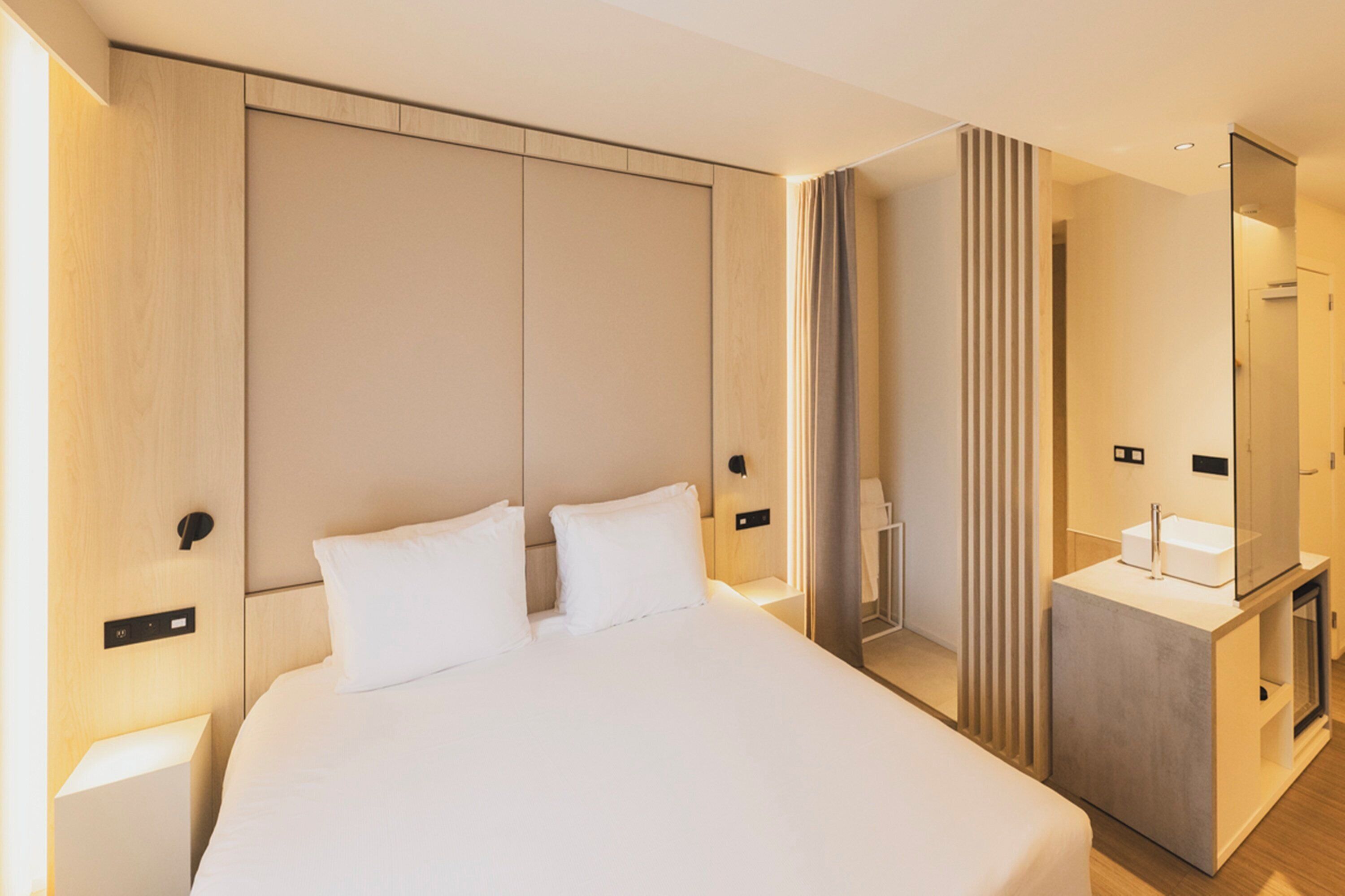Una moderna camera d'albergo con un letto grande, un comodino, una TV integrata e un bagno con specchio e lavandino.