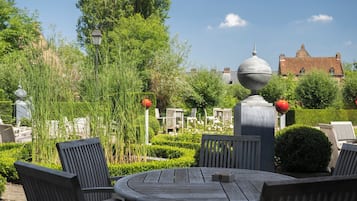 Un giardino con un tavolo rotondo e sedie, circondato da vegetazione e un edificio storico sullo sfondo.