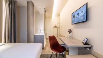 A hotel room with a bed, a desk with a chair, a TV, and a bathroom visible in the background.
