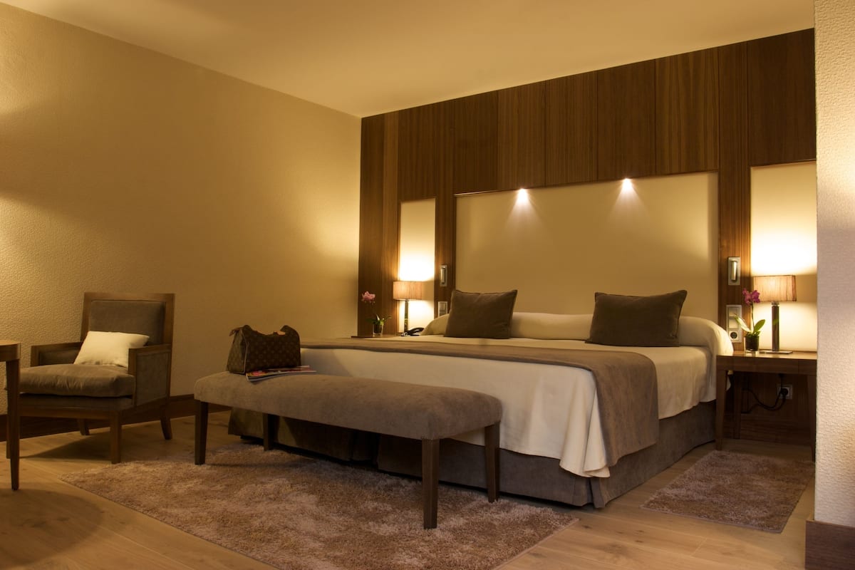 Una habitación de hotel con una cama grande, una silla, un banco y un escritorio con una lámpara.
