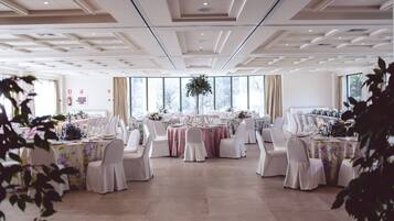 A spacious banquet hall with high ceilings, large windows, and a central floral arrangement. Tables are set with white chairs and floral tablecloths.
