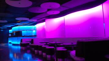 A dimly lit room with round tables and stools, a bar area with blue lighting, and a purple-lit wall.