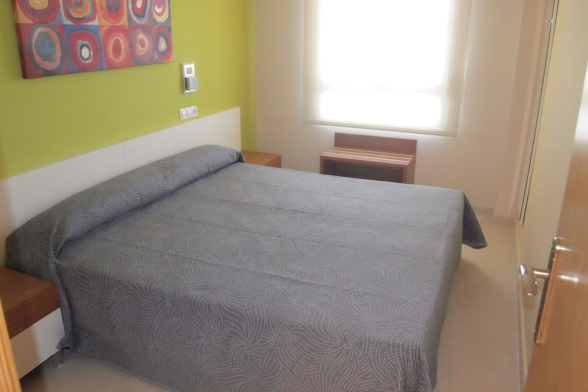 Un dormitorio con una cama, una mesita de noche, un banco de madera, una obra de arte en la pared y una ventana.