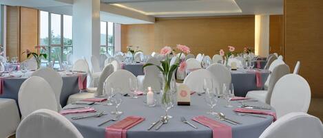 Un salón de banquetes con mesas redondas, sillas blancas y servilletas rosas.