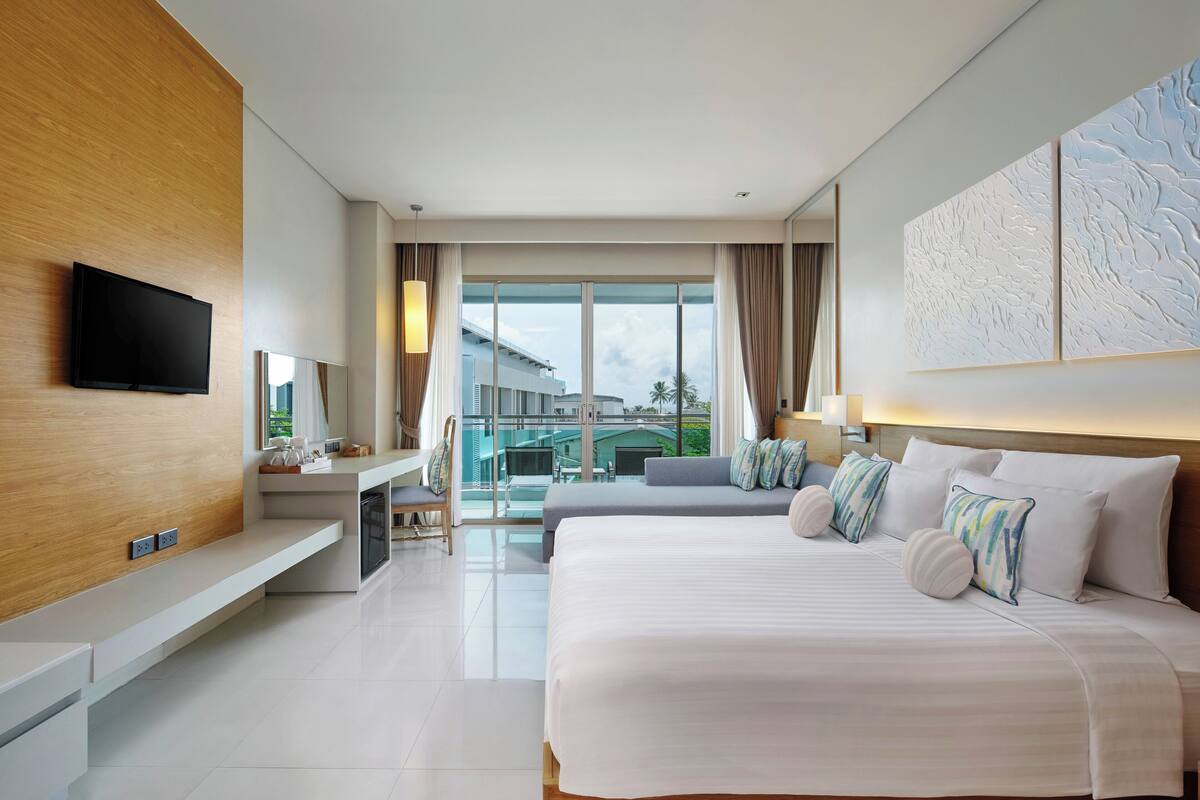 A modern hotel room with a large bed, a flat-screen TV, a desk with a chair, and a balcony with a view of the city.