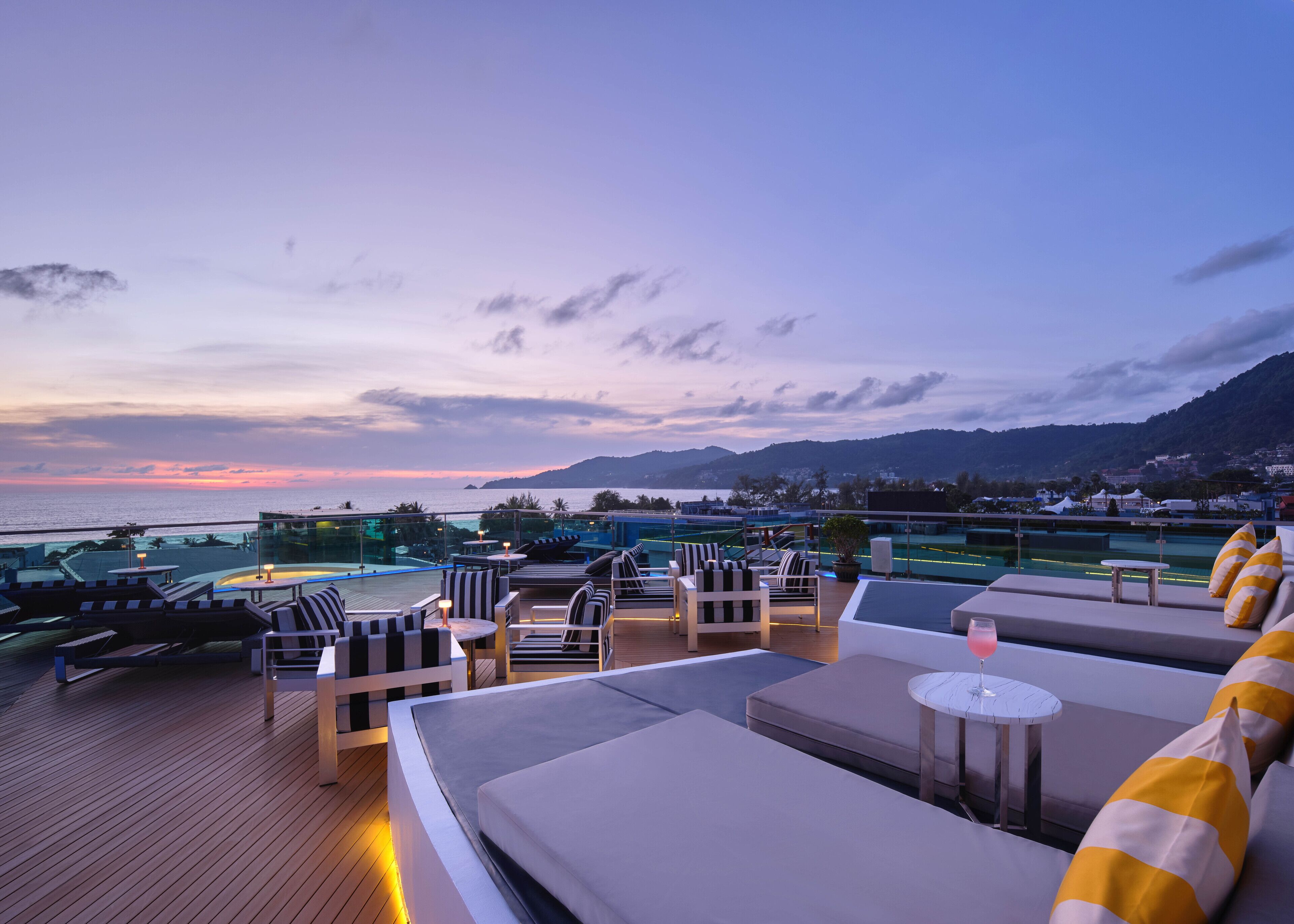 A rooftop lounge with outdoor seating, a pool, and a view of the sea and mountains.
