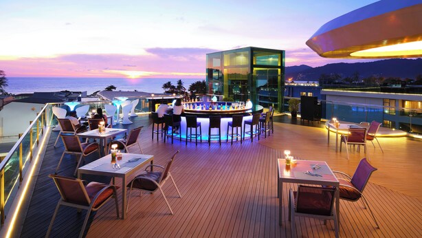 A rooftop bar with a glass enclosure, wooden deck, and seating areas overlooking the ocean at sunset. - The Kee Resort & Spa (Patong)