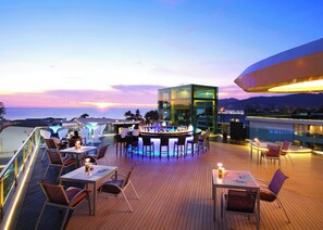 Un bar en la azotea con cerramiento de vidrio, terraza de madera y zonas de estar con vistas al océano al atardecer.