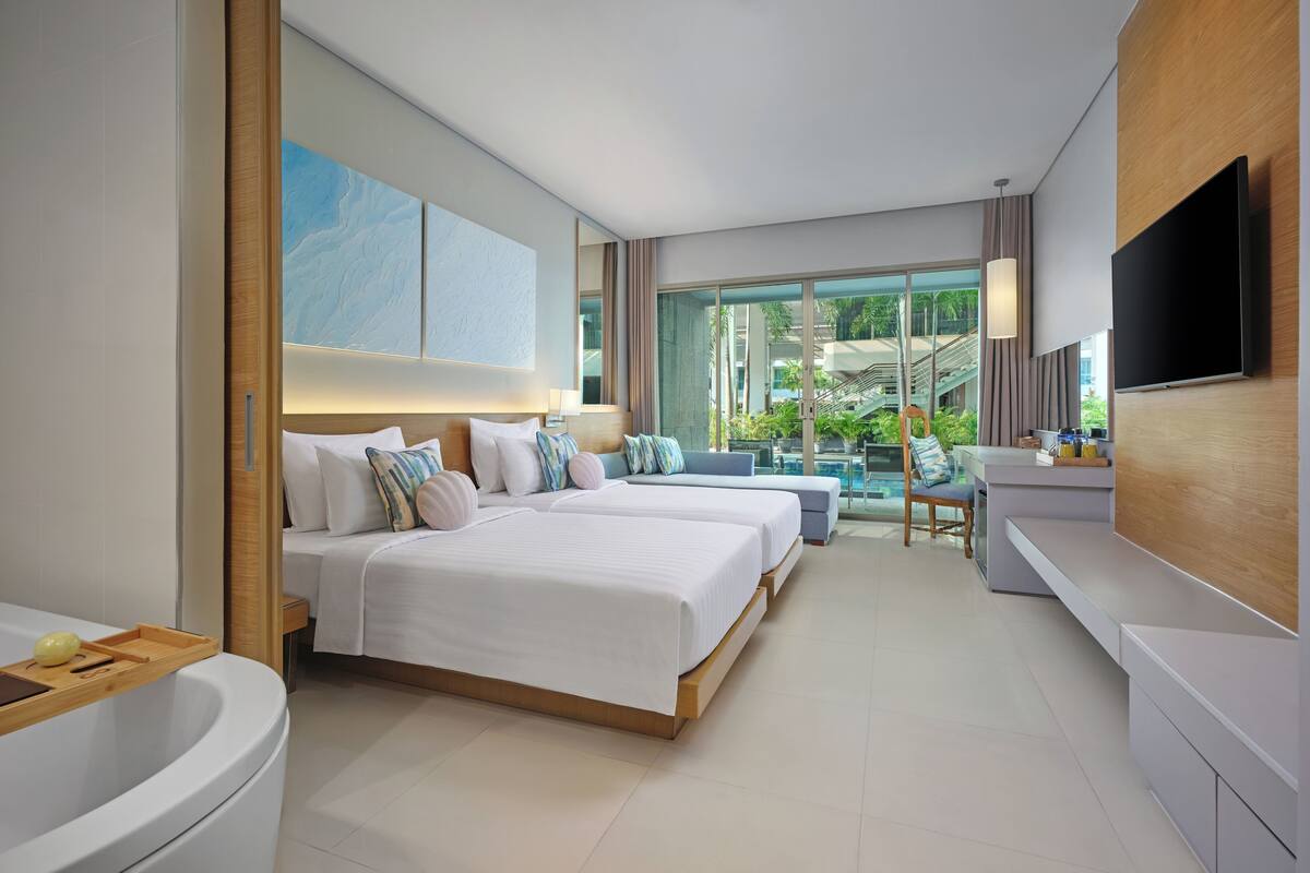 A modern hotel room with two beds, a large TV, a bathroom, and a balcony with a view of the outdoors.