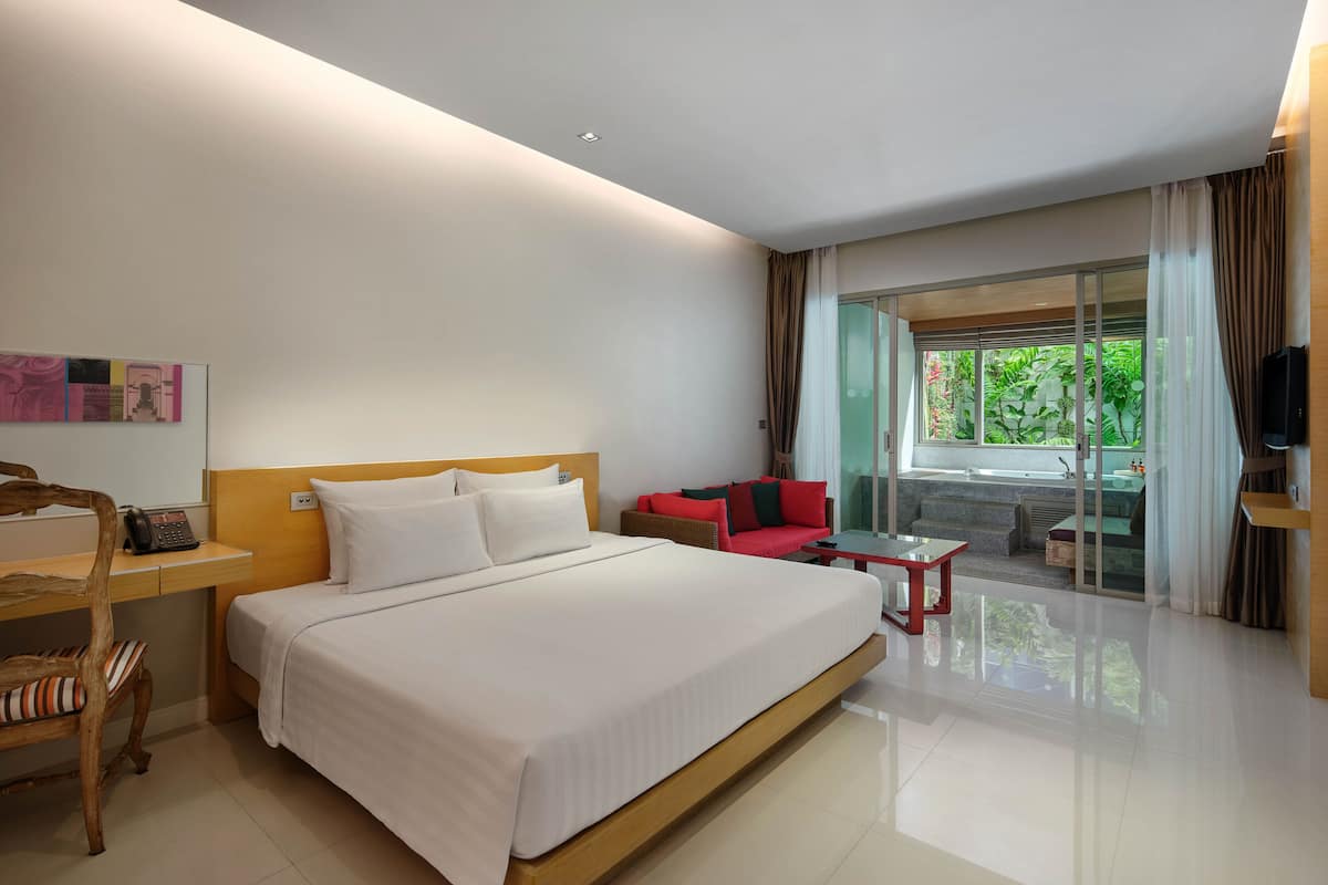 A modern hotel room with a large bed, a desk, a red sofa, and a glass door leading to a balcony with a view of greenery.