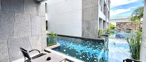 A modern outdoor pool area with a glass table and chairs, surrounded by stone walls and a view of a building in the background.