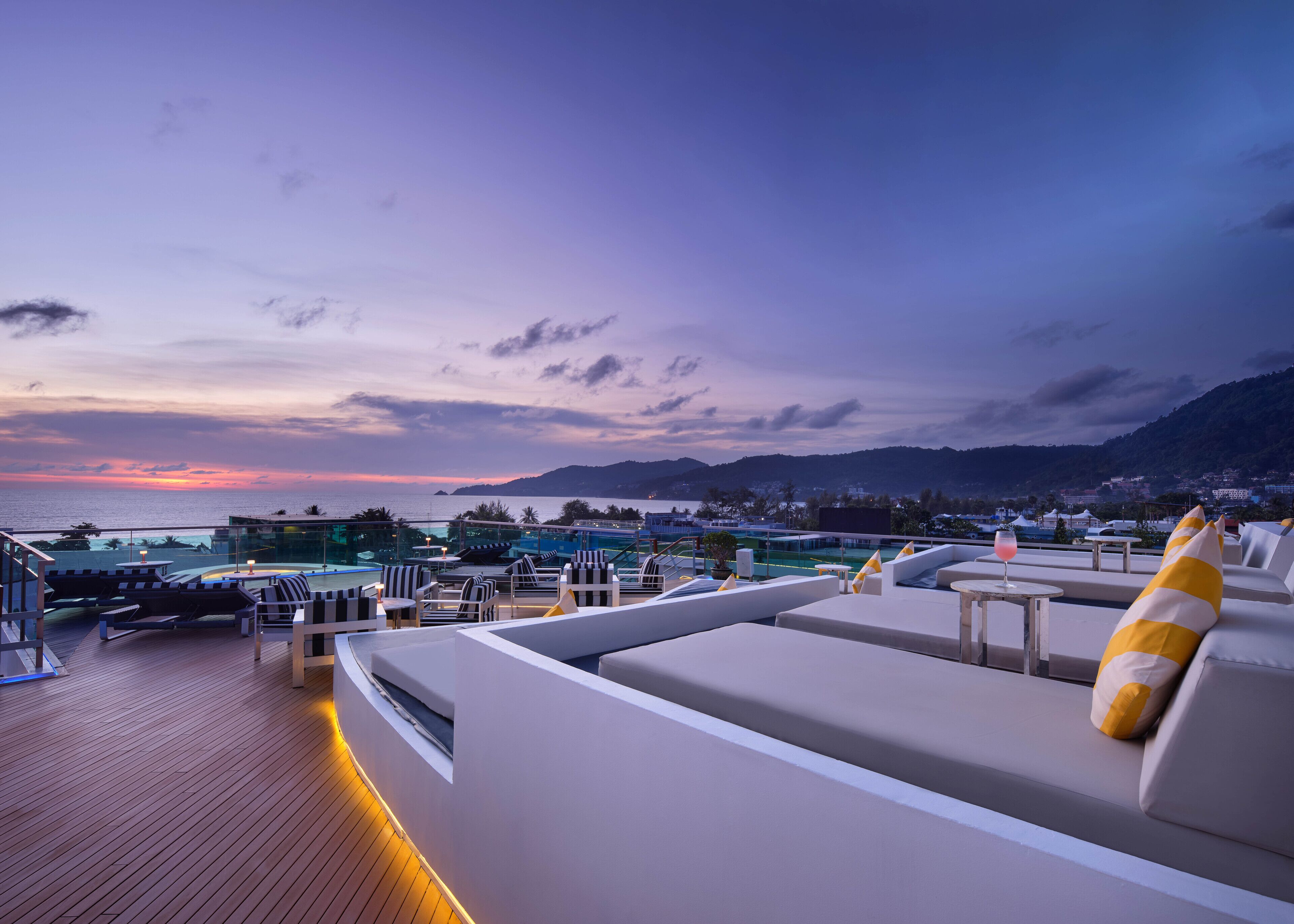 A rooftop lounge with a view of the sunset over the sea, lounge chairs, and a pool area.