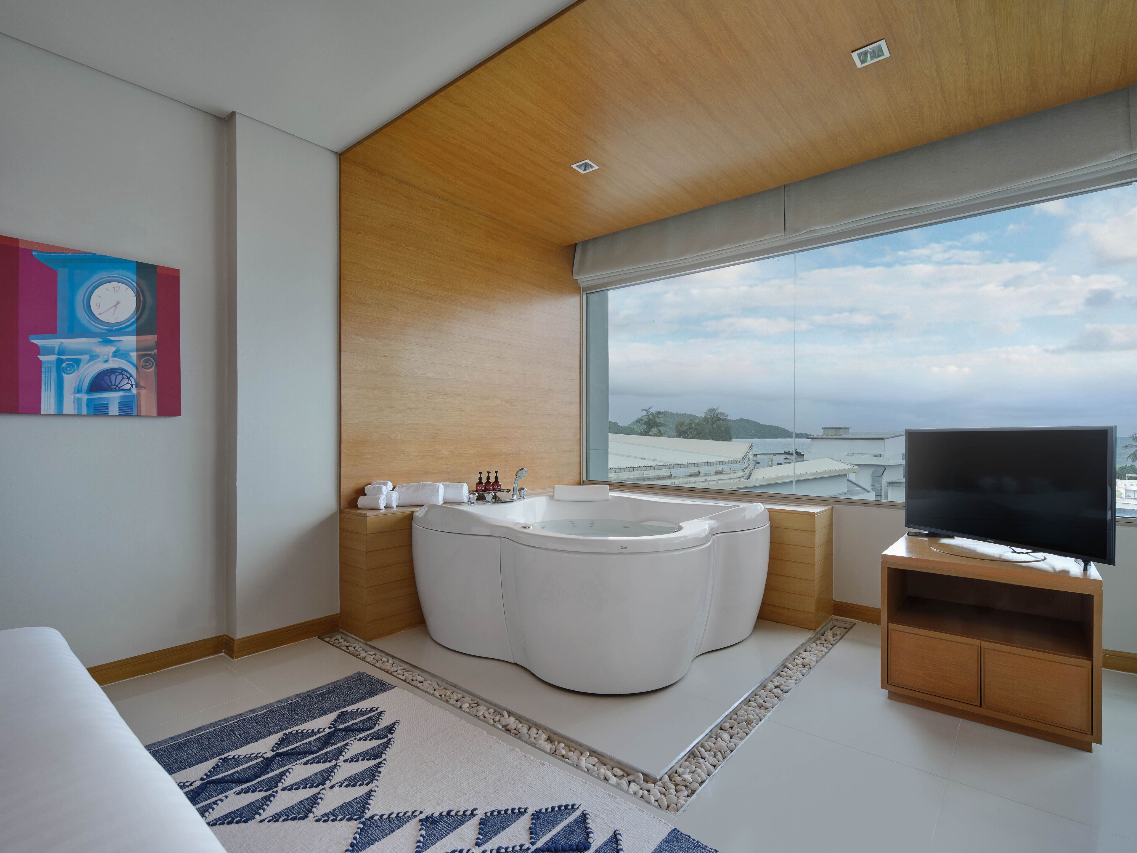 A modern bathroom with a bathtub, a large window, and a television on a wooden stand.