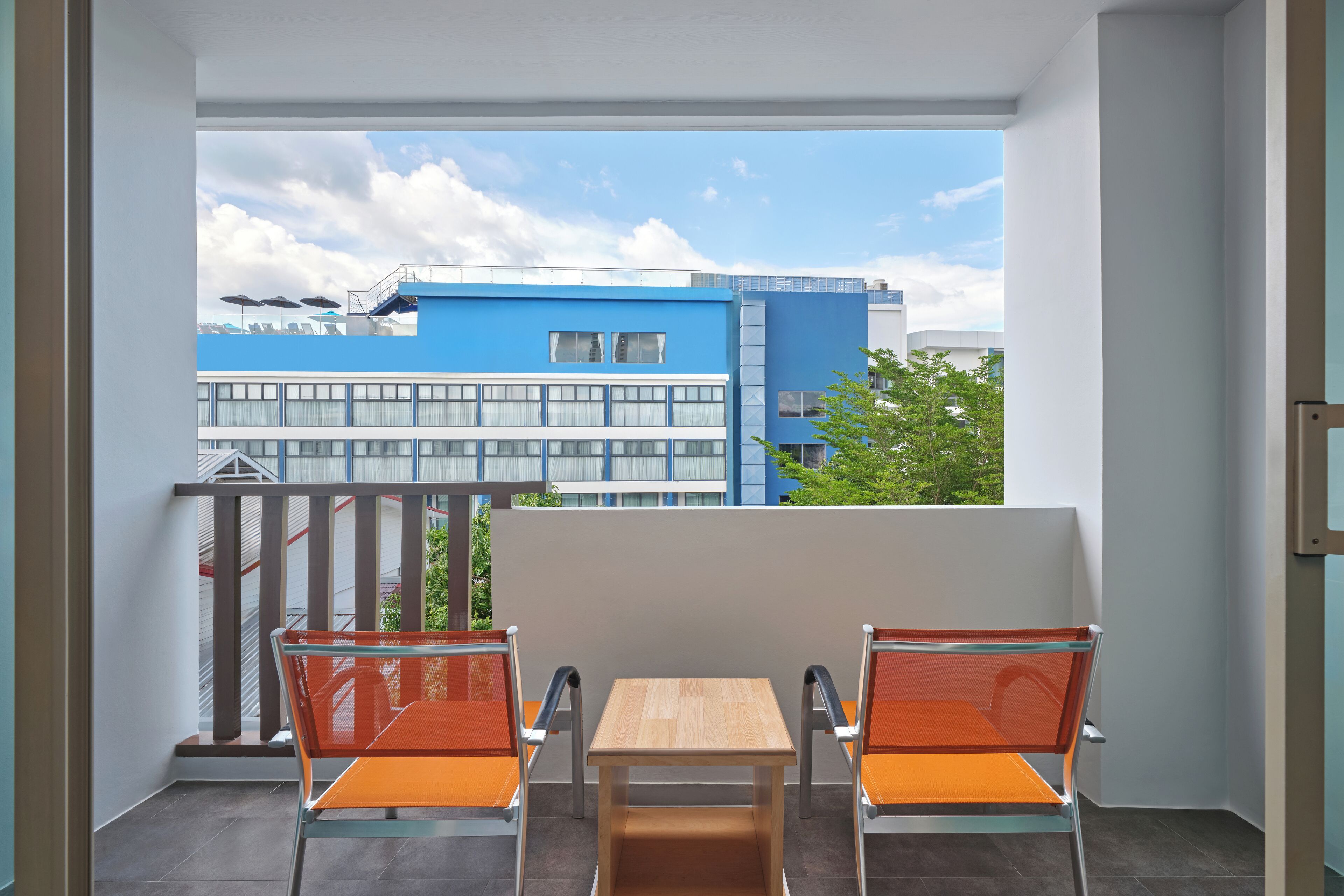 Un balcón con dos sillas naranjas y una mesa de madera, con vistas a un edificio azul con ventanas y un cielo despejado.