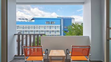 Un balcón con dos sillas naranjas y una mesa de madera, con vistas a un edificio azul con ventanas y un cielo despejado.