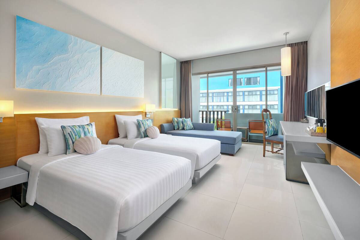 A hotel room with two beds, a desk, a TV, and a balcony with a view of buildings.