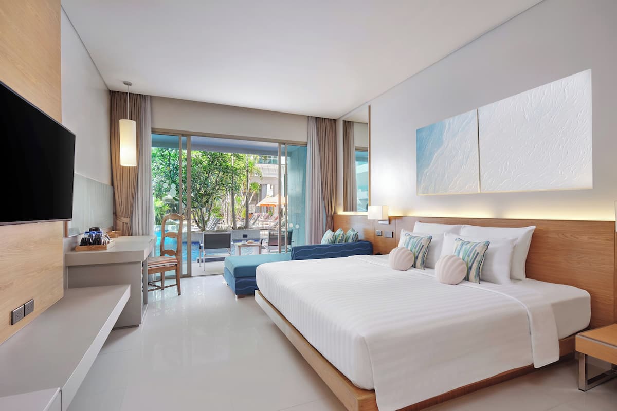 A modern hotel room with a large bed, a desk with a flat-screen TV, a seating area with a pool view, and a balcony with a view of the pool and greenery.