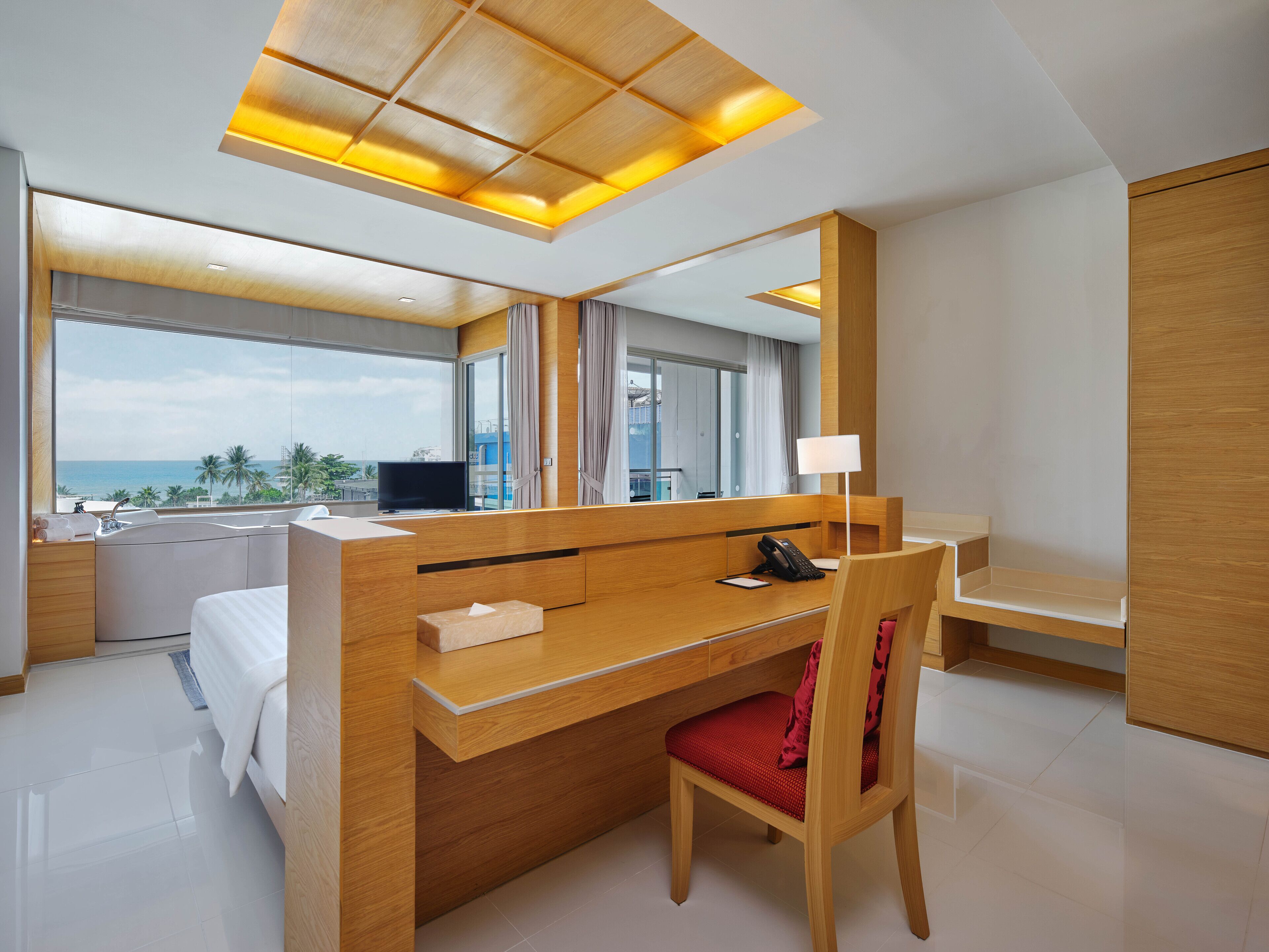 Una habitación de hotel moderna con un escritorio de madera, una silla roja, un espejo grande, un televisor y vista al océano.