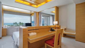 Una habitación de hotel moderna con un escritorio de madera, una silla roja, un espejo grande, un televisor y vista al océano.