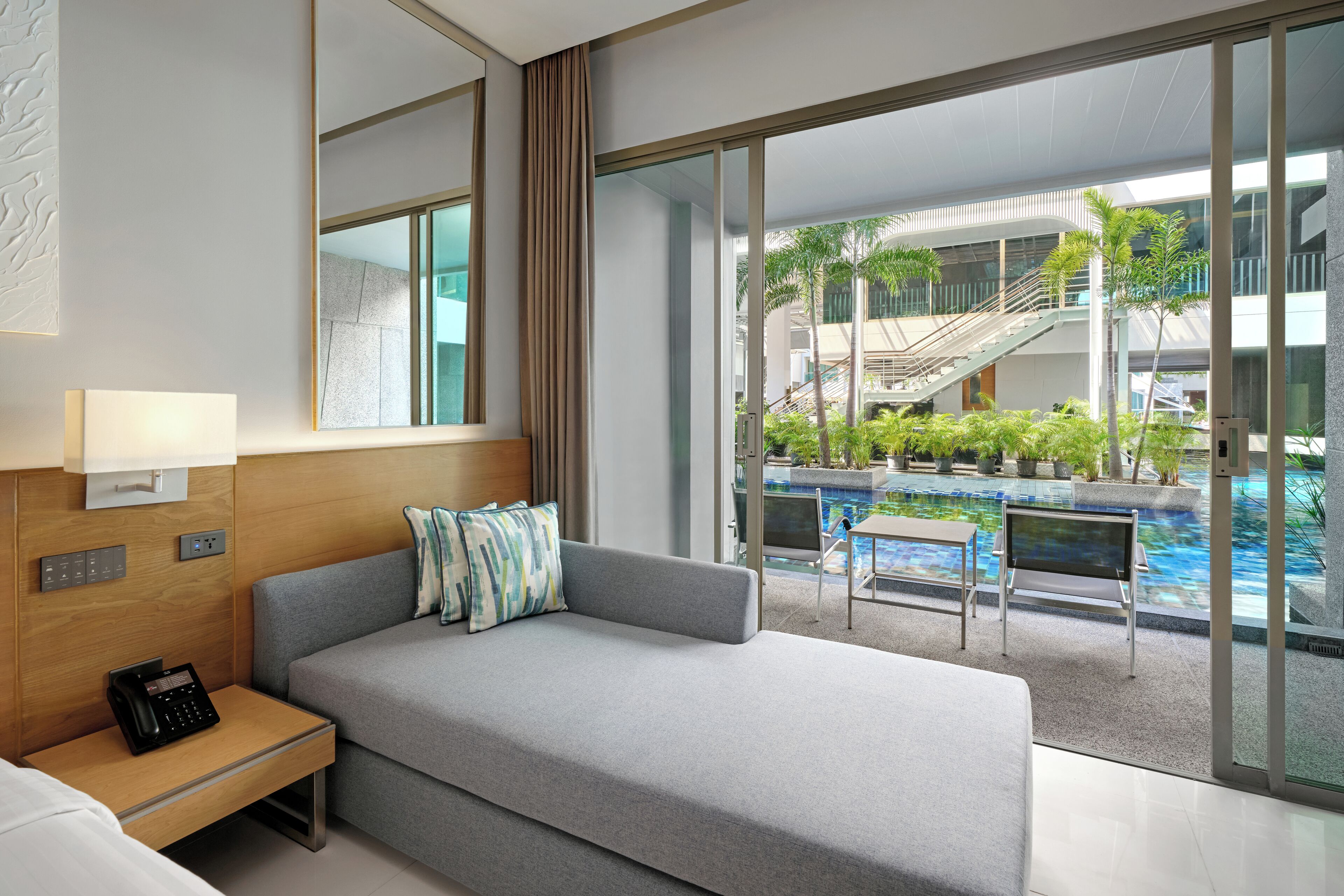 A modern hotel room with a large bed, a bedside table with a phone, a window with a view of an indoor pool and a staircase.
