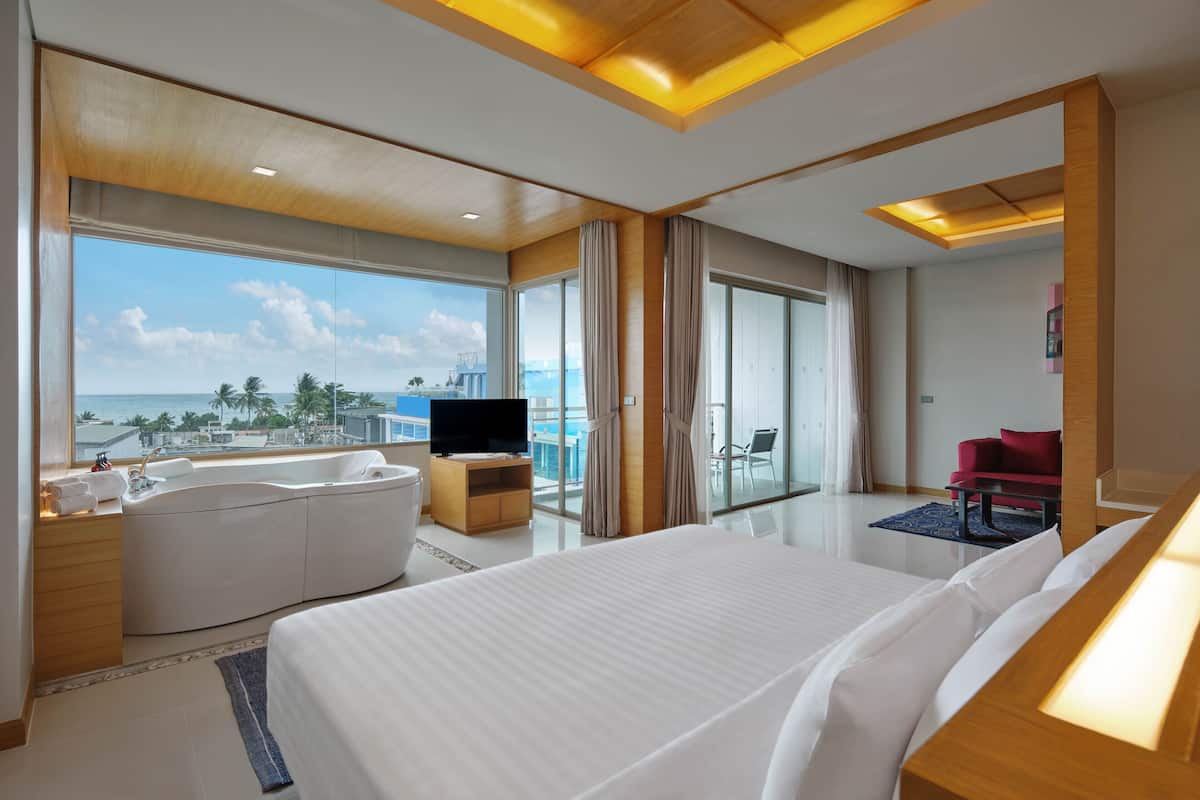 A modern hotel room with a large bed, a TV, a bathtub, and a balcony with a view of the ocean.