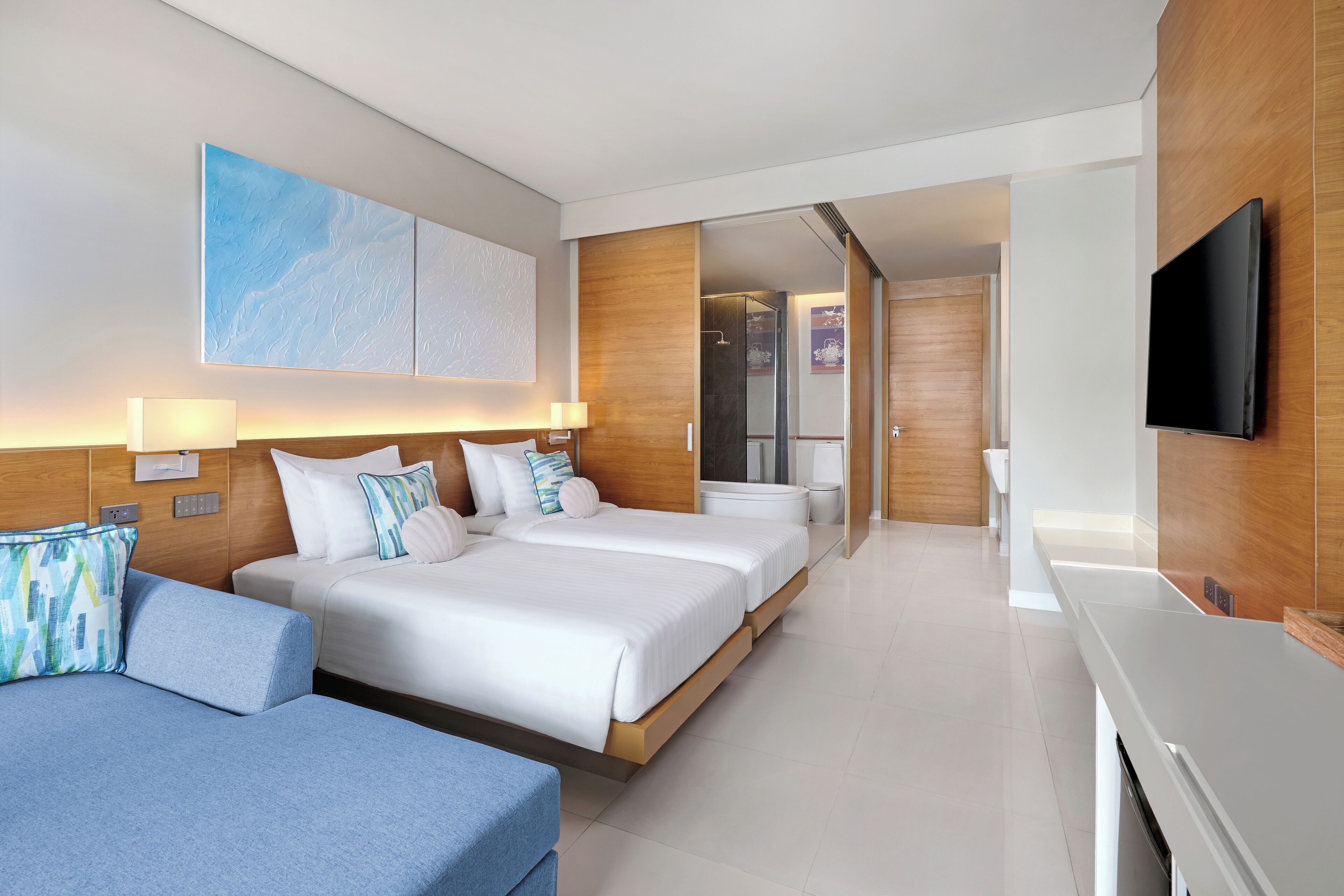 Une chambre d’hôtel moderne, dotée d’un grand lit, d’un canapé, d’une télévision et d’une salle de bain visible à travers une porte ouverte.