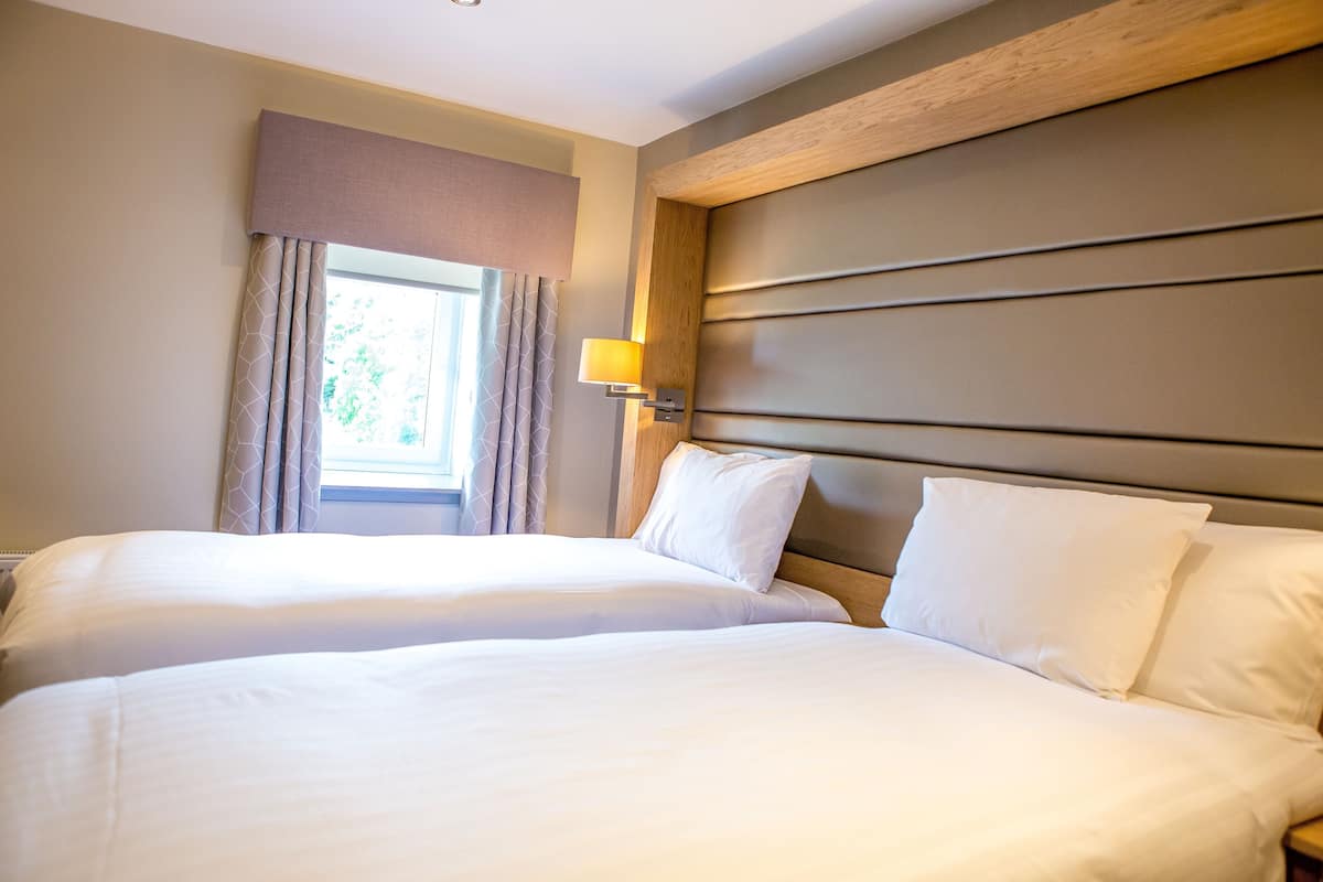 A hotel room with two beds, a window with curtains, a wall-mounted lamp, and a wooden headboard.