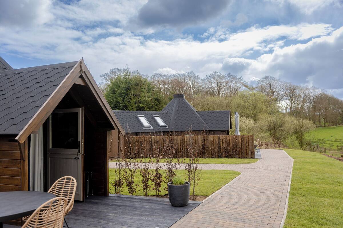 A wooden cabin with a sloped roof, a patio with wicker chairs, and a pathway leading to a grassy area.