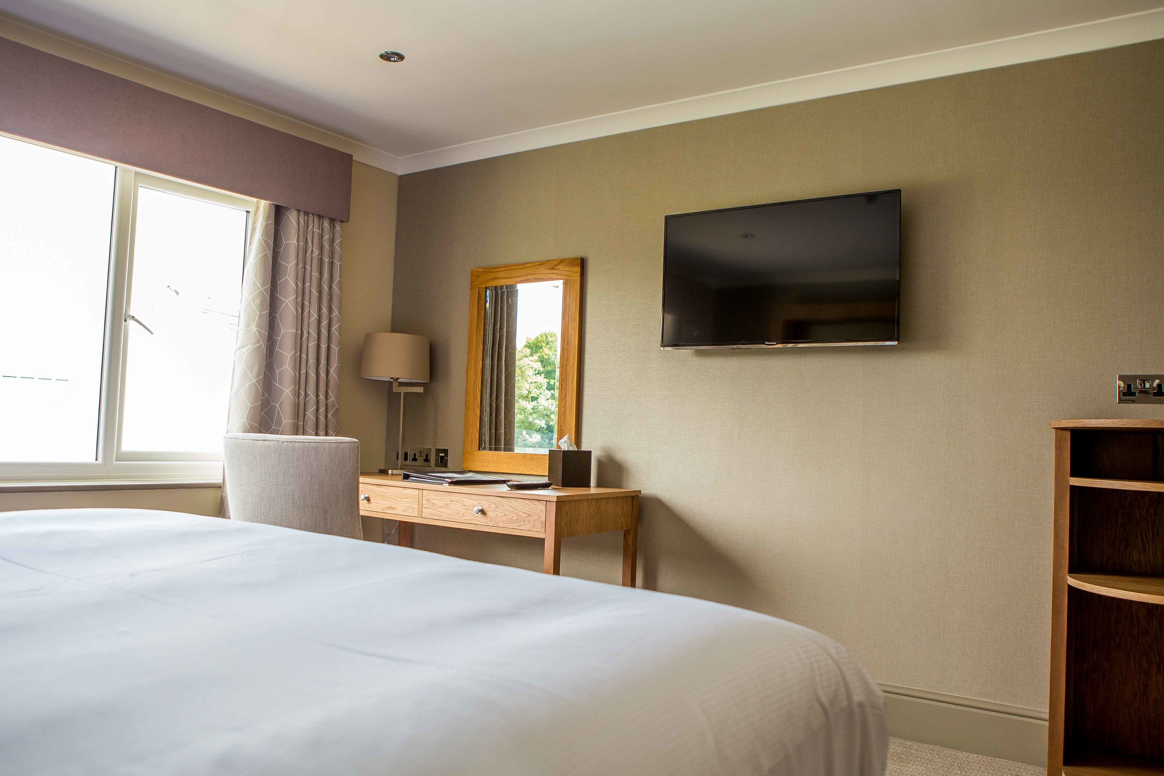 A hotel room with a large bed, a desk with a lamp, a TV mounted on the wall, and a window with curtains.