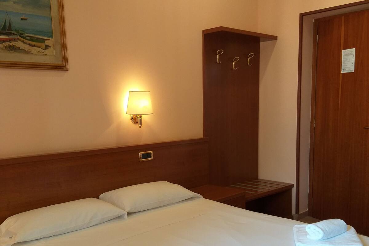 A hotel room with a large bed, a nightstand, a wooden wardrobe, and a wall-mounted lamp.