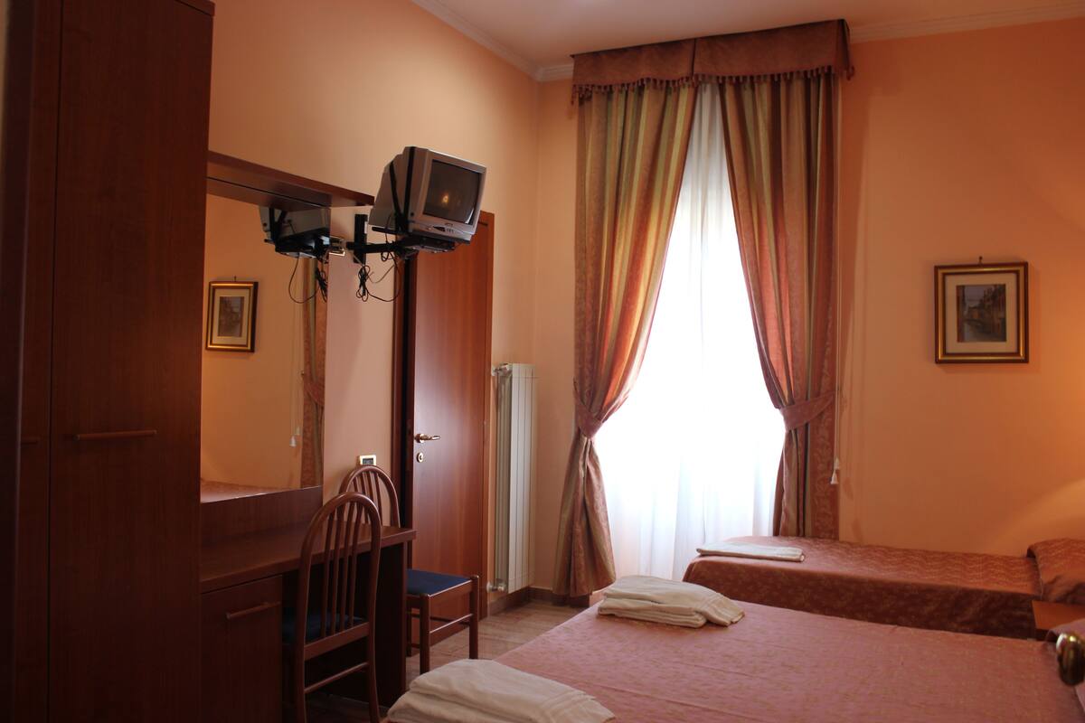 A hotel room with a bed, a desk with a chair, a television mounted on the wall, and a window with curtains.
