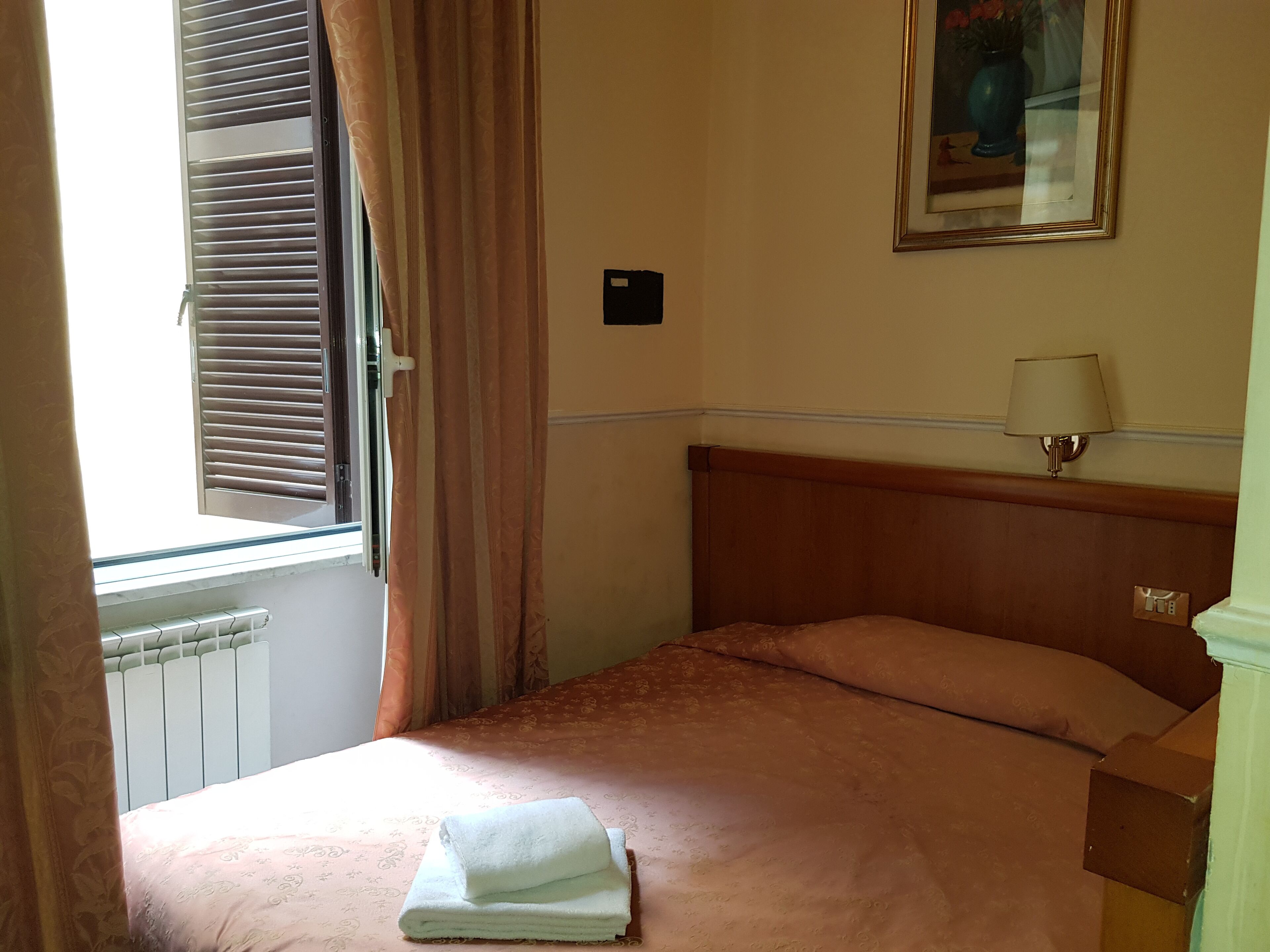 Una habitación de hotel con una cama, una ventana con persianas, un radiador, una lámpara de pared y un cuadro enmarcado.