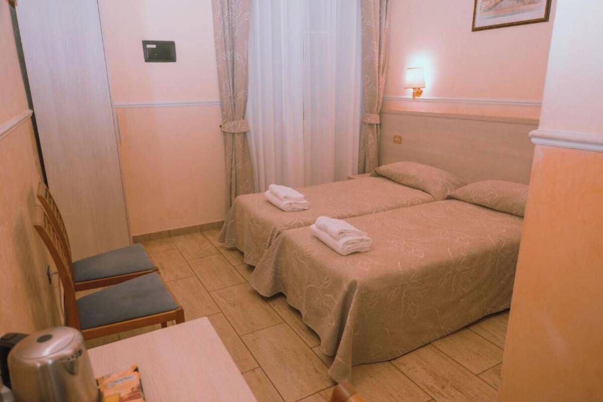 A hotel room with two beds, a table with a kettle, and a chair.