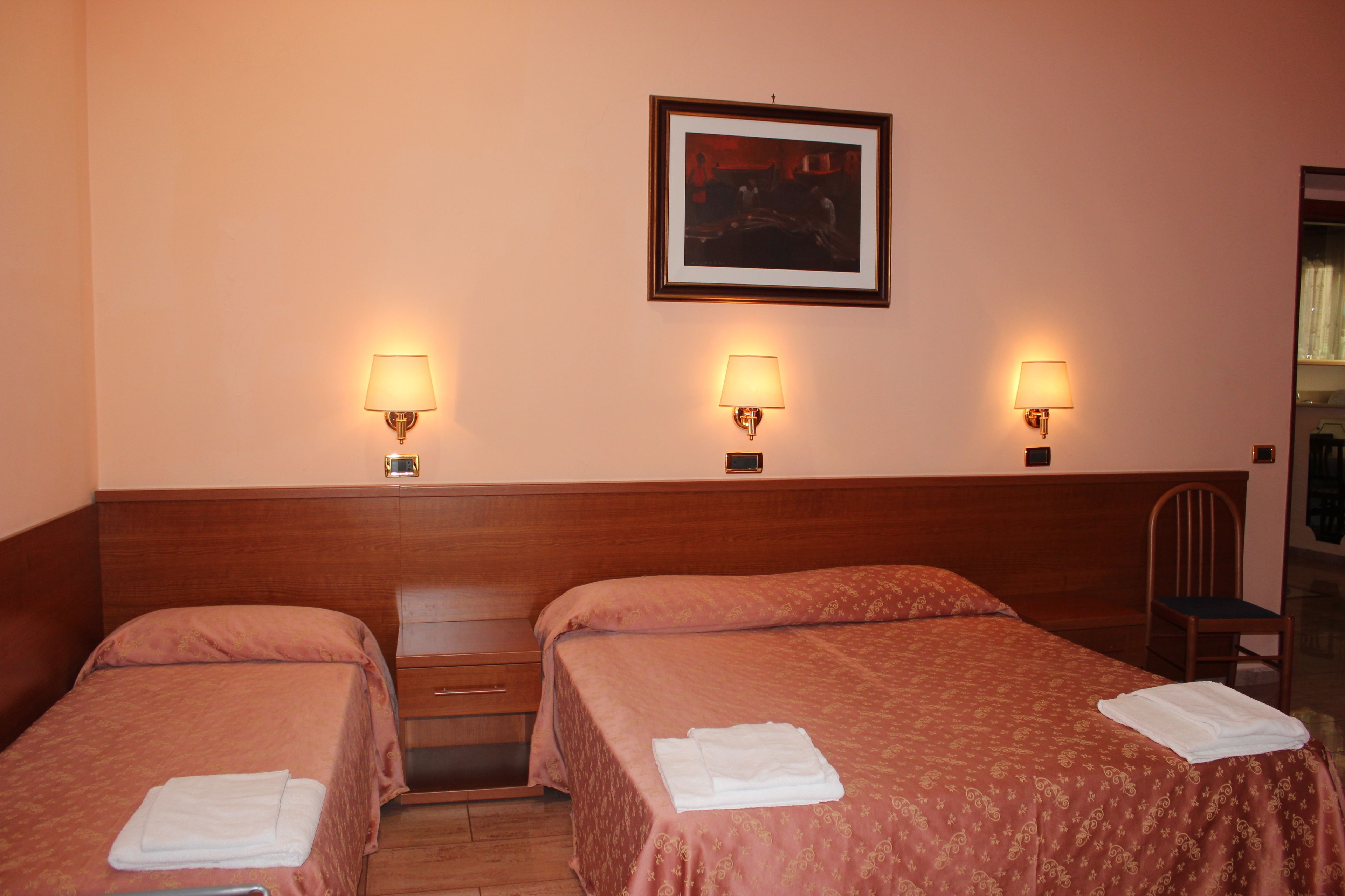 Habitación de hotel con dos camas, cada una con colcha roja y toallas blancas. Hay tres lámparas de pared y un cuadro enmarcado sobre las camas.