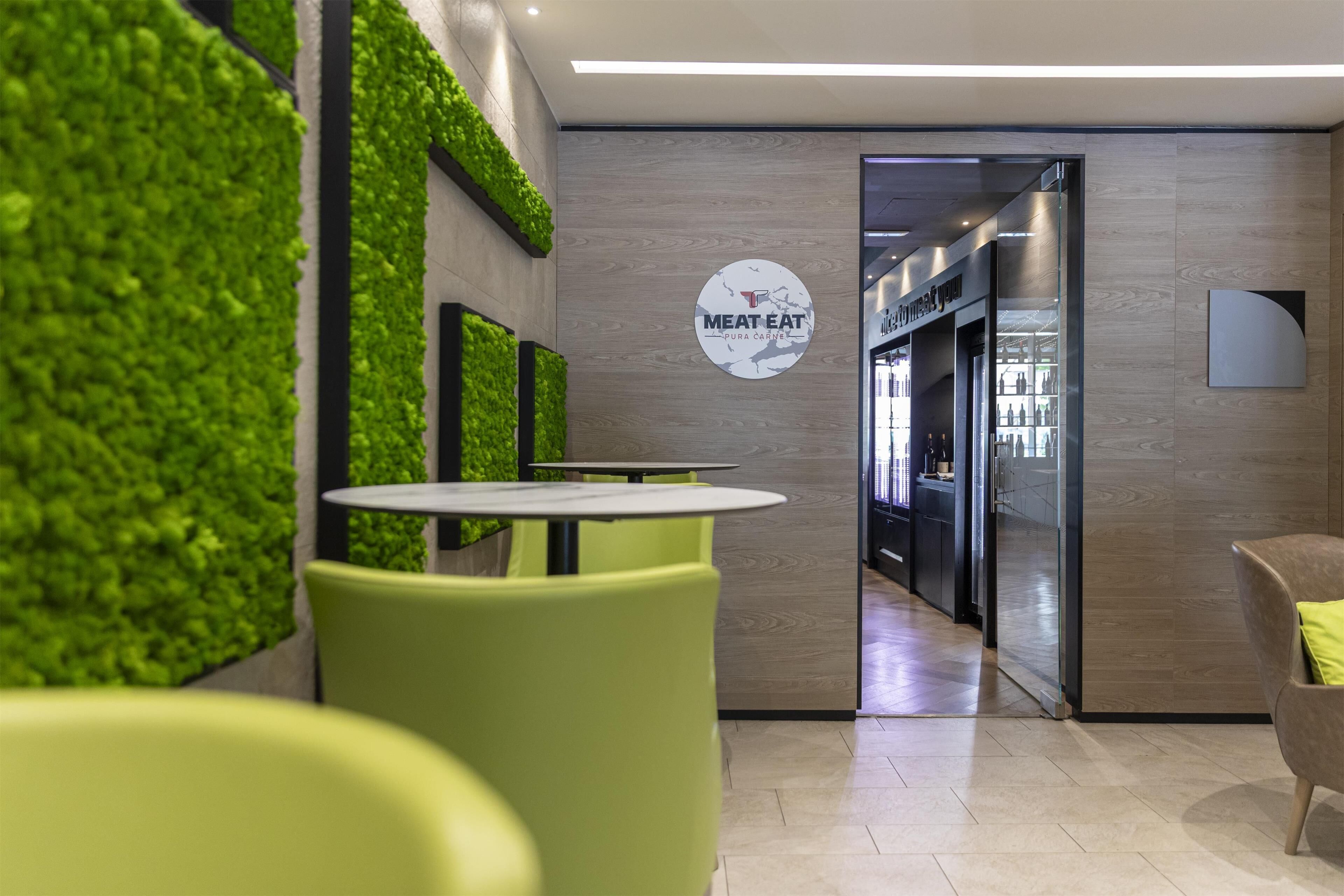 L’intérieur d’un restaurant moderne, avec des sièges verts, un tableau des menus bien visible et une cuisine en arrière-plan.