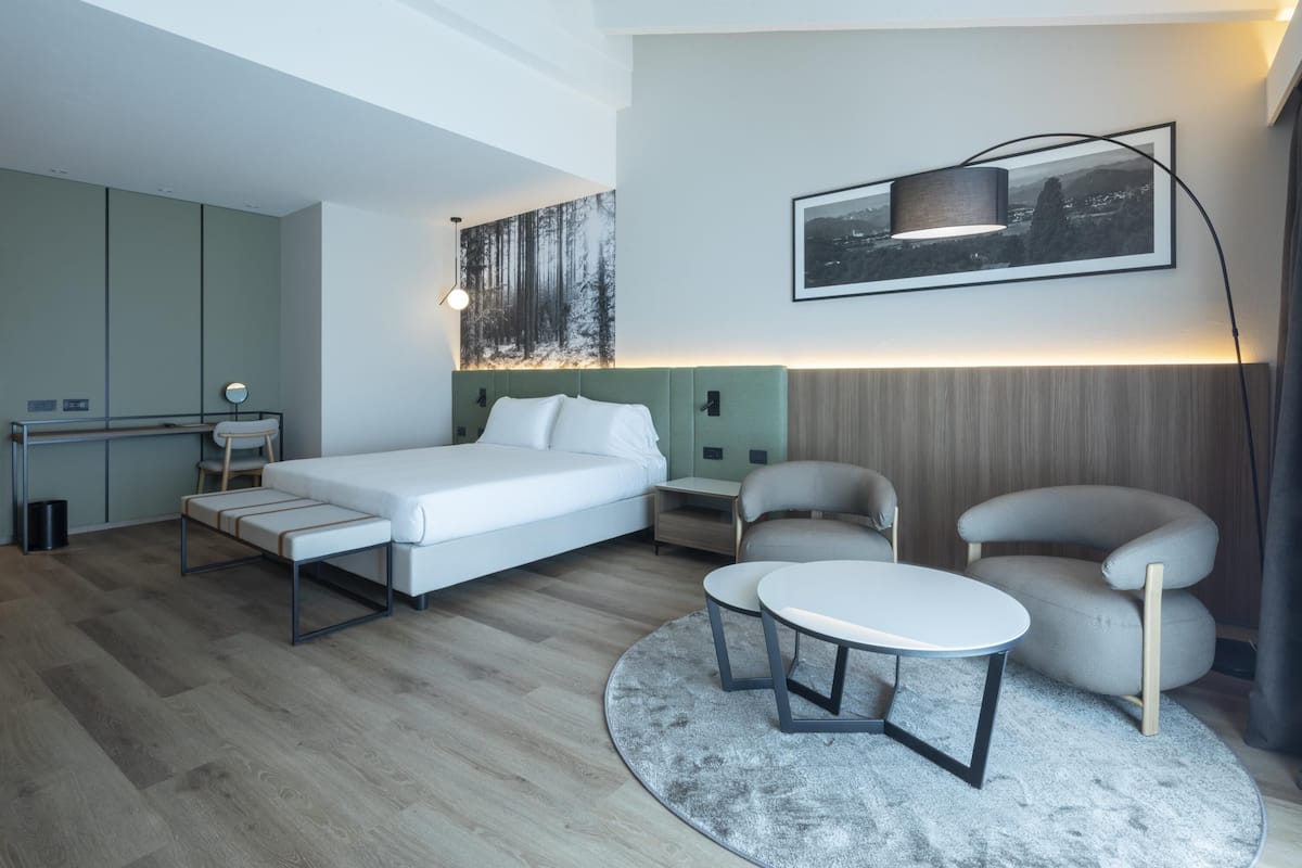 A modern hotel room with a bed, a desk, a chair, a round table, and a painting on the wall.