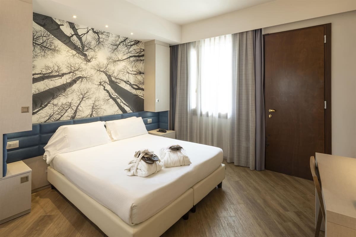 A hotel room with a large bed, a nightstand, a desk, and a wardrobe.