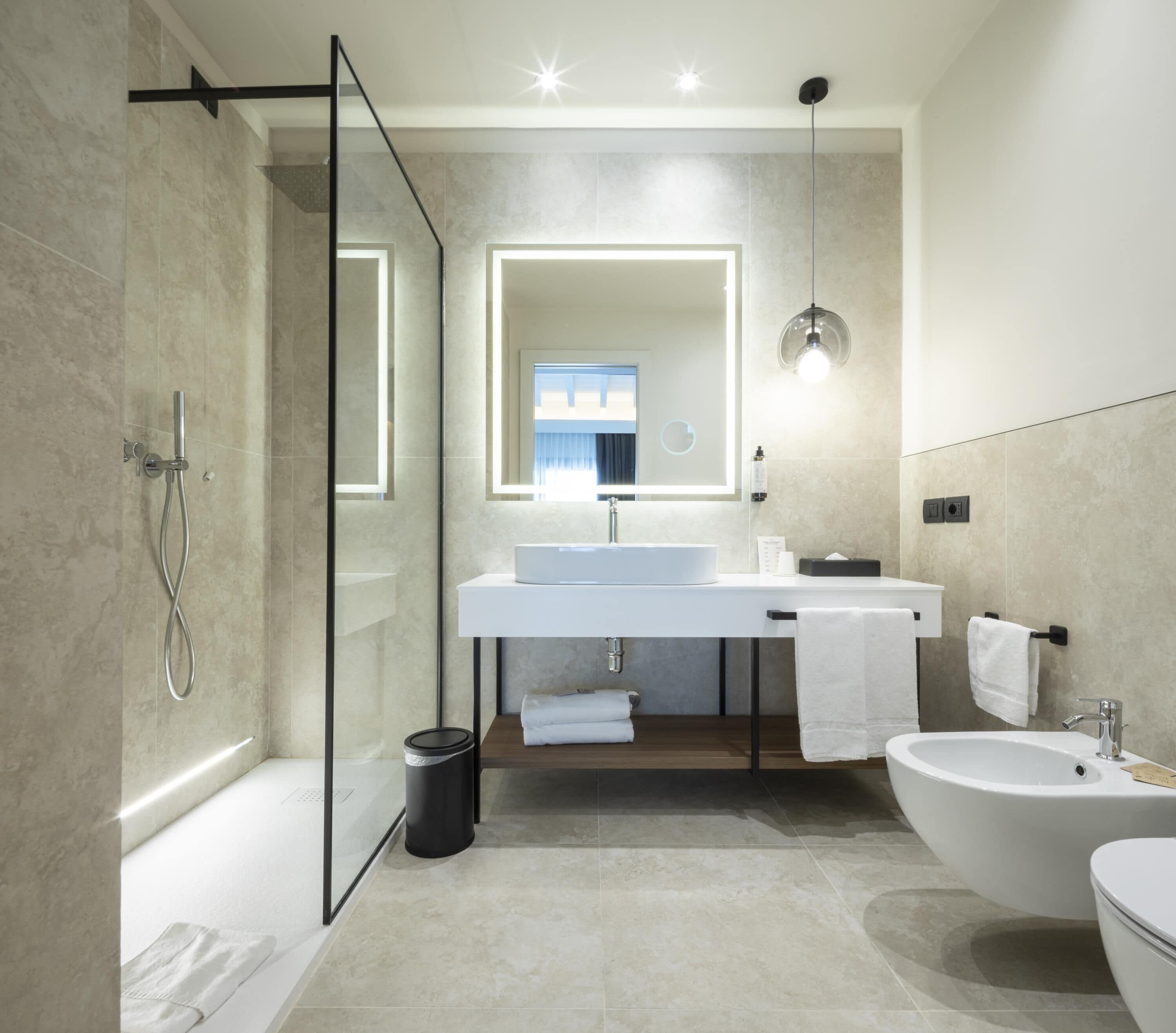 A modern bathroom with a glass shower enclosure, a large mirror, a white sink, a bidet, and a toilet.