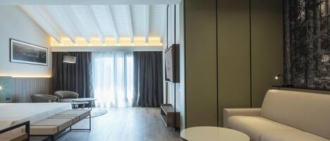 A modern hotel room with a sofa, a dining table, a TV, and a large window with curtains.