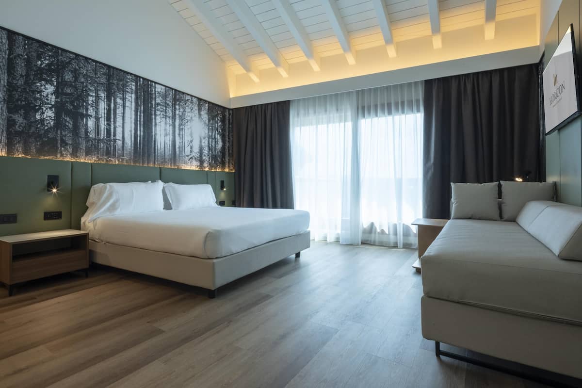A modern hotel room with a large bed, a sofa, a nightstand, a wall-mounted TV, and a large window with curtains.