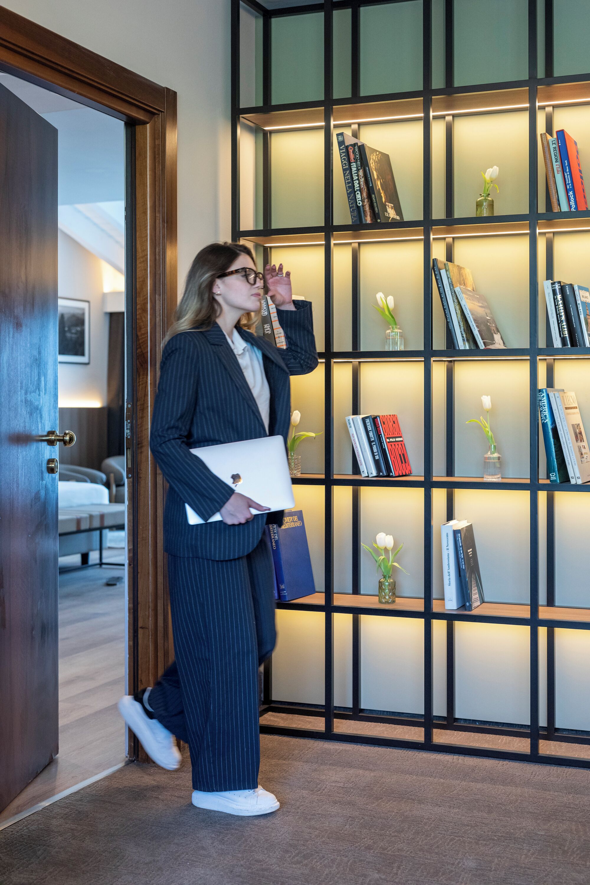 A person in business attire standing in front of a bookshelf with books and vases.